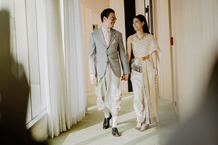 Mew Nittha walking with her partner in a cream-and-gold Chakkraphat outfit with a shoulder drape and full-length skirt.
